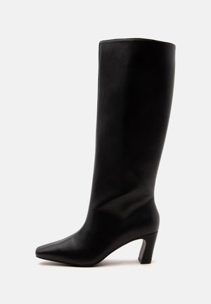 Botte en cuir noire montée jusqu'au genou avec un bout pointu et un talon bloc de hauteur moyenne sur un fond clair uni.