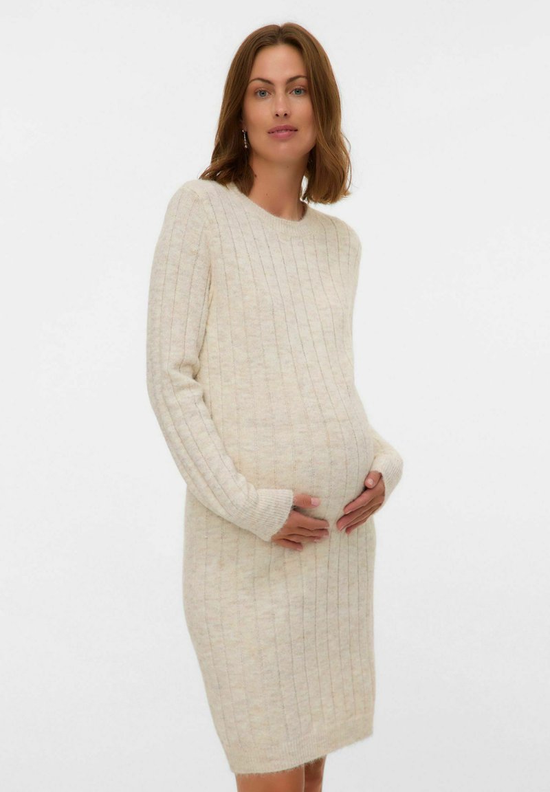 Rased naine püsivalt beige ribikoes kleidis õrnalt oma kõhtu hoides, seistes ühetaolise valge tausta ees.