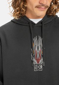 Sudadera negra con un gráfico de llamas en blanco, rojo y naranja, con un logo de DC impreso debajo. El material parece suave y texturizado.