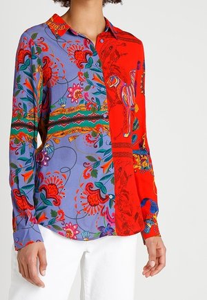 Camisa de manga larga con botones y estampados florales llamativos en púrpura y rojo, con diseños coloridos de caballos y hojas, combinada con pantalones blancos.