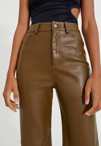 Pantalons en simili cuir marron avec une texture lisse, taille haute, poches avant et couture visible le long des coutures. Détail de bouton argenté inclus.