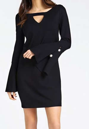 Mujer vistiendo un minivestido negro de manga larga con mangas acampanadas y detalles de botones plateados, con un pequeño corte en el escote en forma de ojo de cerradura.