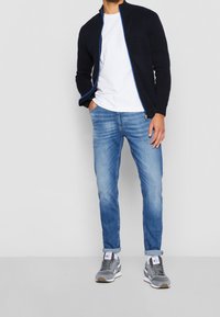 Marinblå dragkedjejacka med blå accenter, vit t-shirt, ljusblå jeans och grå sneakers med vita och bruna sulor.