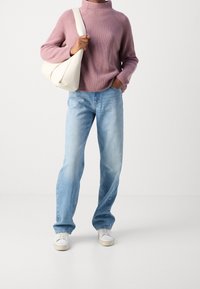 Light purple ribbed sweater, blue denim jeans, and white sneakers. The model holds a cream shoulder bag, standing against a plain background.