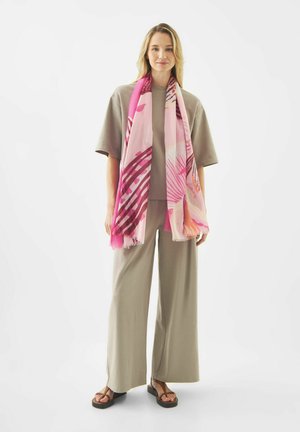 Vrouw draagt taupekleurige losse broek en T-shirt, staat met een roze- en paarspatroon sjaal om haar nek gedrapeerd, en zwarte sandalen.