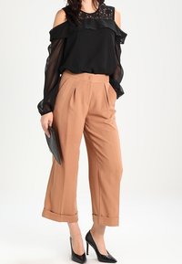 Blusa nera senza spalle con dettagli in pizzo, abbinata a pantaloni plissettati in tinta beige a vita alta e tacchi a punta neri. Tiene una clutch nera.