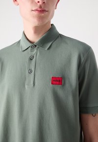 Polo-shirt i olivengrønn, laget av bomull, med en ribbestrikket krage, tre knappelukkinger, og et rødt HUGO-merke på brystet.