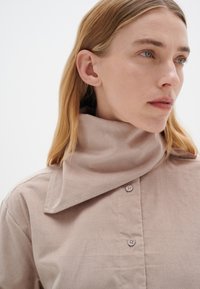 Lysebrunt kordfløjlsskjorte med en høj, foldet krave og to synlige knapper. Stoffet har en tekstureret, ribbet overflade.