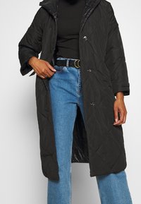 Abrigo largo acolchado negro con capucha, que presenta grandes botones, bolsillos laterales y un corte relajado, llevado sobre una camiseta negra y jeans azules.