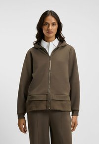 Brun zip-up hoodie med mjuk pälskant längst ner. Har en huva och en avslappnad silhuett. Bärs över en vit skjorta med krage.