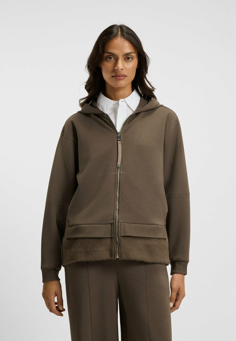 Brun zip-up hoodie med mjuk pälskant längst ner. Har en huva och en avslappnad silhuett. Bärs över en vit skjorta med krage.