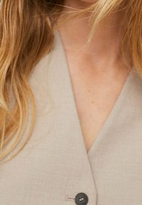 Gilet in tessuto beige leggero con scollo a V, caratterizzato da un singolo bottone nero sul davanti e una texture sottile.