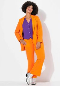 Completo arancione con giacca aderente e pantaloni a zampa, abbinato a un gilet viola con bottoni. Il modello indossa scarpe in metallo argento.