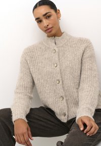 Licht beige gebreide cardigan met een hoge kraag, zeven grote knopen en een zachte, gestructureerde oppervlakte. Lange mouwen met ribgebreide manchetten.