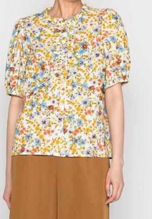 Kvinde iført en kortærmet blomstret bluse med flæser og knapper, kombineret med brune bukser med høj talje.