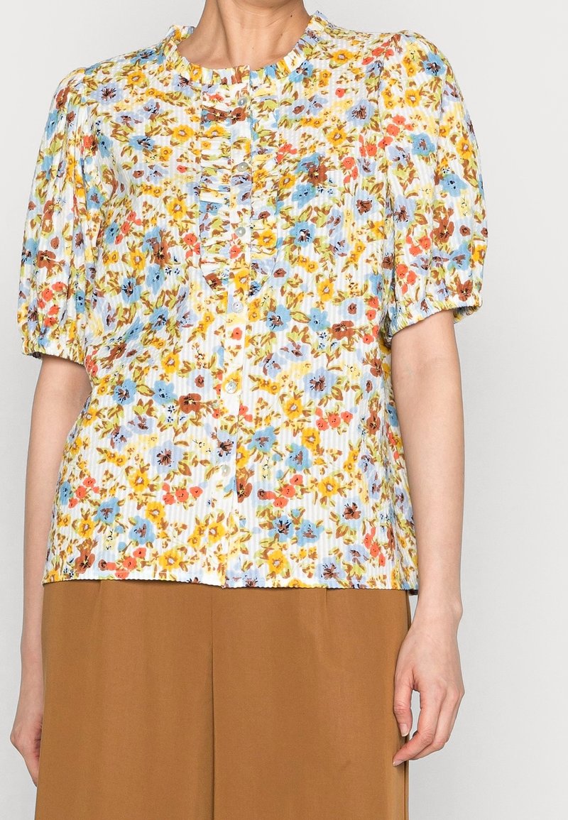 Femme portant un chemisier à manches courtes à fleurs avec des volants et des boutons, associé à un pantalon marron taille haute.