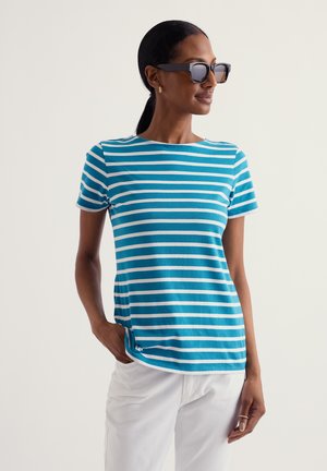Femme portant des lunettes de soleil noires, un chemisier à manches courtes rayé bleu et blanc, un pantalon blanc, la main dans la poche, regardant sur le côté.