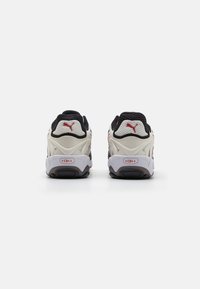 Black and beige athletic shoes with a textured mesh upper, thick white midsole, and black rubber outsole. Red logo accents on heel.