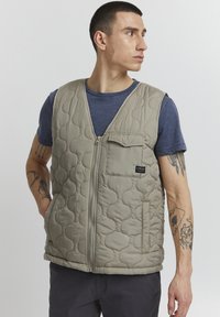 Quilted, beige padded vest with a V-neck, front zipper, and a chest pocket. Features textured pattern and a logo tag on the pocket.