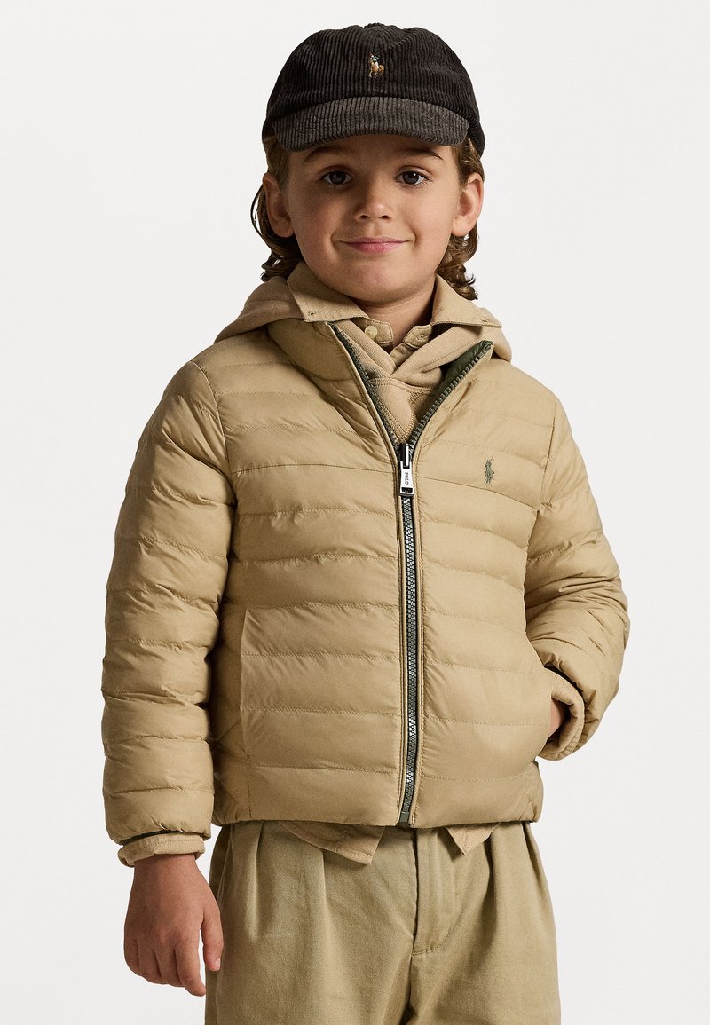 Béžová puff jacket s zipsom, rebrovanými manžetami a malým logom. Nosí sa cez béžovú mikinu a svetlo béžové nohavice, doplnené tmavou čiapkou.