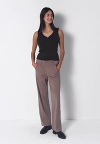 Femme aux longs cheveux foncés portant un haut noir sans manches, un pantalon large taupe et des chaussures noires, debout devant un fond blanc.