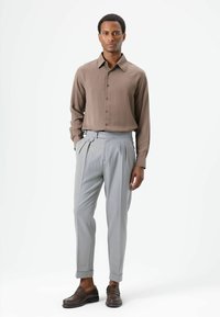 Chemise marron à manches longues avec boutons, pantalon gris clair fuselé et mocassins marron. Tissu lisse et coupe ajustée avec détails plissés.