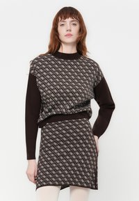 Donna con capelli rossi che indossa un maglione a fantasia marrone e una gonna abbinata, in piedi contro uno sfondo bianco con una mano sulla vita.