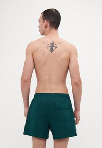 Shorts de bain verts avec une ceinture élastique et une poche arrière, en tissu texturé. Le modèle est montré de dos.
