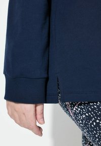 Camisa de manga larga en color navy con puños acanalados y aberturas laterales, exhibida junto a leggings estampados con un diseño multicolor.