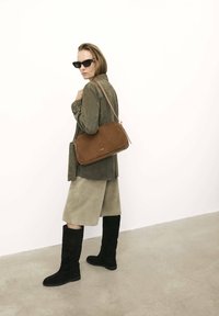 Borsa a tracolla in pelle marrone con forma rettangolare e superficie liscia. Il modello indossa una giacca in suede verde e una gonna beige, stivali neri fino al ginocchio.