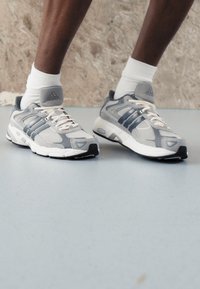 Sive i bijele atletske cipele sa mrežastim gornjim dijelom, tri plave pruge i bijelim vezicama. Imaju strukturalnu đon s crnim akcentima.