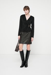 Black long-sleeve wrap top with a decorative gold accent, paired with a dark leather skirt and knee-high black boots. Small brown bag included.