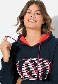 Felpa blu navy con fodera interna rossa e logo decorato. Presenta un cappuccio con cordino dai toni viola e rosa. Texture liscia, design casual.
