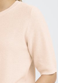 Īsām piedurknēm, gaiši rozā adīta blūze ar apaļu kakla izgriezumu, teksturētu adījuma rakstu un piegulošu siluetu. Šuvju detaļas ir minimālas.