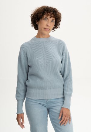 Femme aux cheveux courts et bouclés portant un pull en tricot bleu clair et un jean bleu clair, debout devant un fond blanc uni.