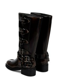 Bottes en cuir synthétique noir arrivant au genou, dotées de plusieurs sangles à boucles argentées, d'un bout arrondi et d'un talon noir épais.