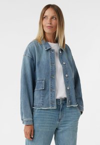 Kobieta o włosach blond do ramion, ubrana w jasnoniebieską kurtkę dżinsową i dżinsy, z białą bluzką pod spodem, patrzy lekko na bok.