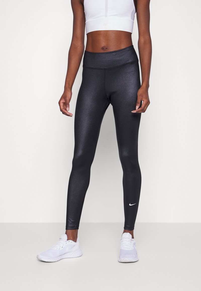 Femme portant des leggings de sport noirs brillants, un débardeur court blanc et des chaussures de course blanches, se tenant devant un fond uni.