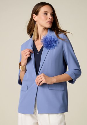 Frau trägt einen hellblauen Blazer mit einer großen blauen Stoffblume als Anstecker, weiße Hose, ein goldenes Armreif und lange goldene Ohrringe, blickt nach rechts.