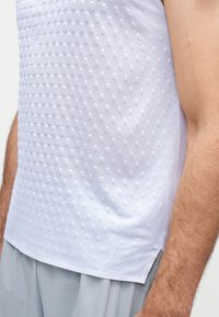 Gros plan d'une personne portant une chemise blanche texturée sans manches avec des motifs en points en relief et un pantalon gris clair.