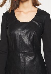 Kobieta w czarnej sukience z długim rękawem, z przednim panelem o skórzanej teksturze i okrągłym dekoltem, odkrywająca część szyi i włosów.