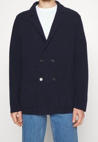 Námořnický pletený cardigan s dvouřadou řadou knoflíků, texturovanou tkaninou, dvěma předními kapsami a černými knoflíky. Nosený přes bílou košili.