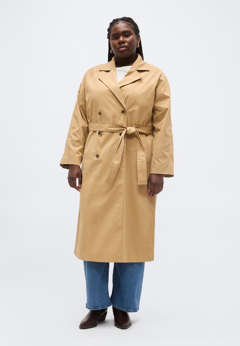 Femme aux cheveux foncés tressés, portant un trench-coat beige ceinturé, un jean bleu et des bottines marron foncé, se tenant devant un fond uni.
