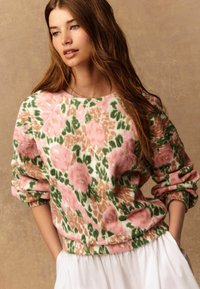Junge Frau mit langen braunen Haaren, die einen rosa, grünen und beigefarbenen, blumig gemusterten Pullover und einen weißen Rock vor einem beigen Hintergrund trägt.