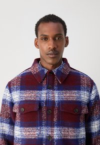Red and blue plaid flannel shirt with a collared neck, two chest pockets, and black button closures. Soft texture with a warm appearance.