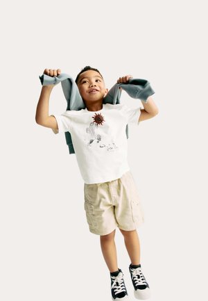 T-shirt blanc avec une impression de cactus et de soleil, short beige et baskets noires. L'enfant tient un sweat-shirt gris sur les épaules, souriant.