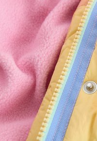 Tejido de polar en un suave color rosa, superpuesto con una tela amarilla que presenta un cierre de cremallera multicolor y un botón de presión plateado. Se aprecian contrastes de textura.