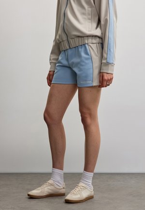 Person, die beige und hellblaue Sportshorts, beige Zip-up-Jacke mit blauem Streifen, weiße Socken und beige Sneaker trägt und auf grauem Boden steht.