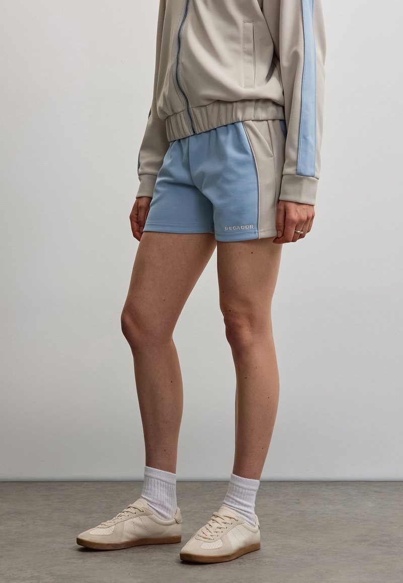 Personne portant un short de sport beige et bleu clair, une veste beige zippée avec une bande bleue, des chaussettes blanches et des baskets beiges debout sur un sol gris.