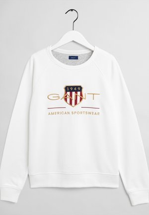 Sweat-shirt blanc à manches longues avec col rond, patch logo indiquant "1949 GANT" et texte "American Sportswear" sur le devant, suspendu sur un cintre blanc.